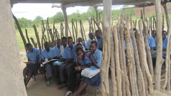 Schulkinder in ihrer alten Schule, die aus in den Boden gesteckten Baumstämmen und einen provisorischem Dach besteht.